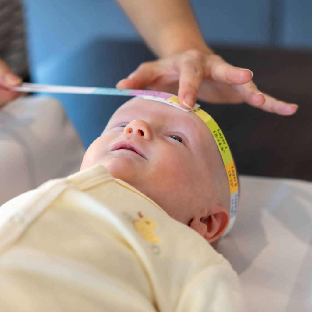 Baby lying on a surface with a measuring tape around its head, held by an adult.