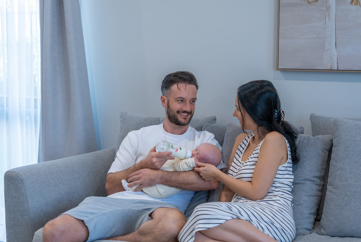 A mum and dad on the couch, smiling at their baby in his dad's arms