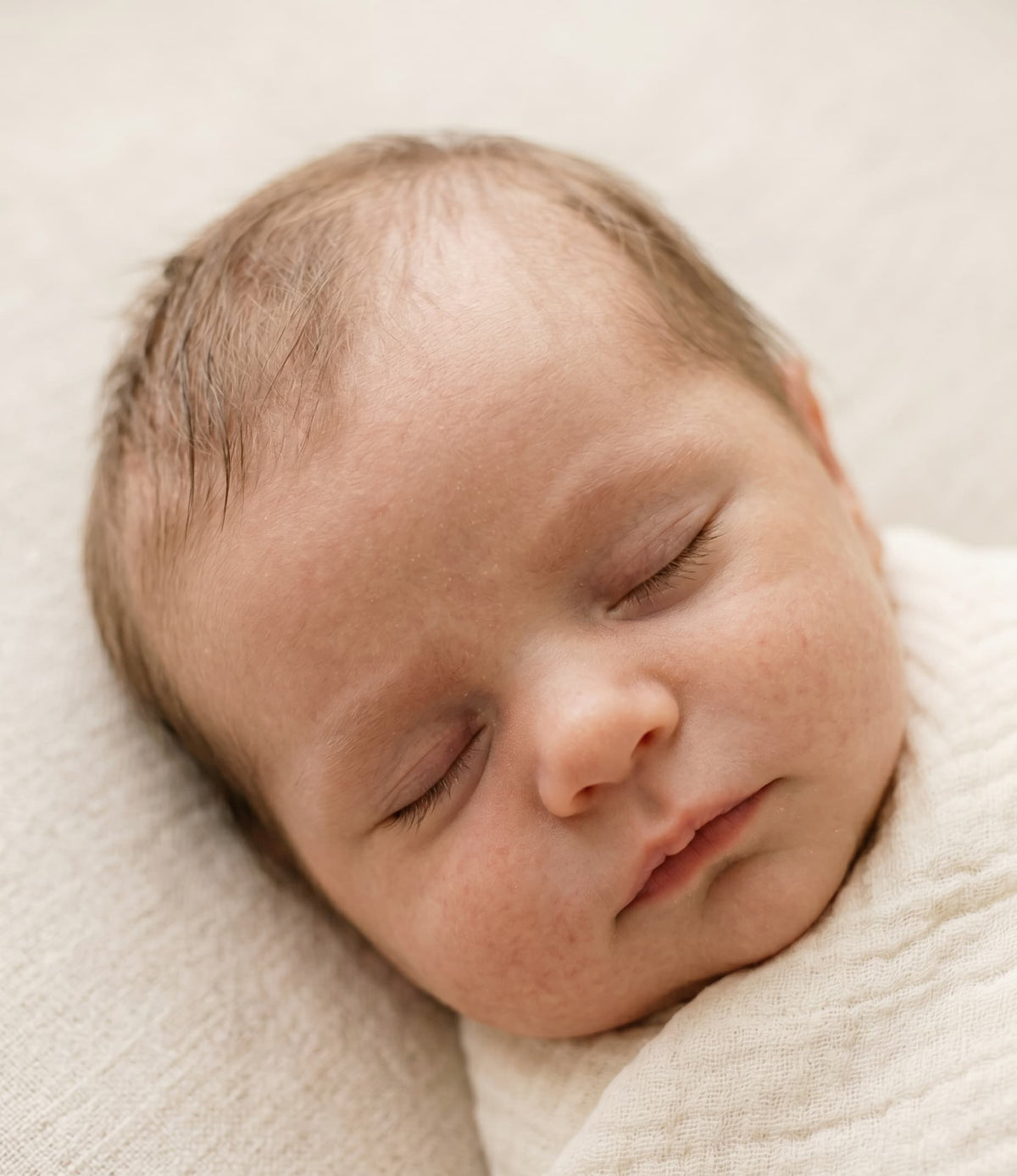 A newborn baby asleep, wrapped in a cream muslin
