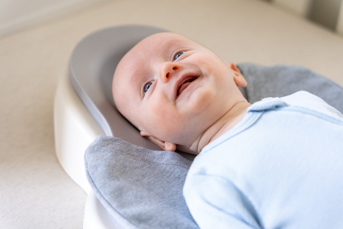 A smiling baby lying on his back on the Perfect Noggin in a cot