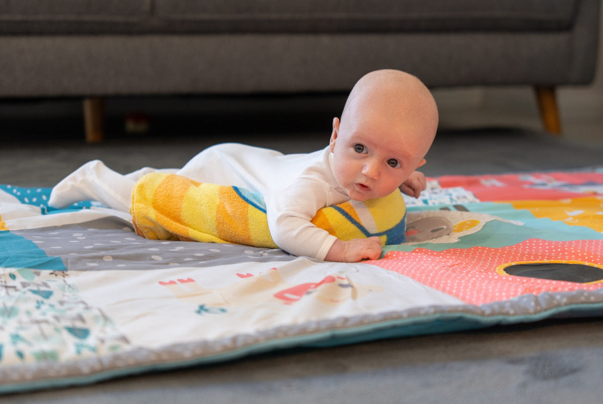 A baby doing tummy time over a rolled towel on a colourful play mat, head up and looking at the camera