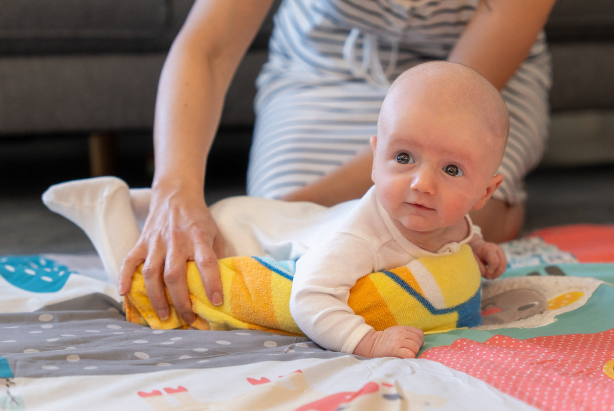A mum steadying a rolled towel under her baby's chest as he lifts his head on a play mat